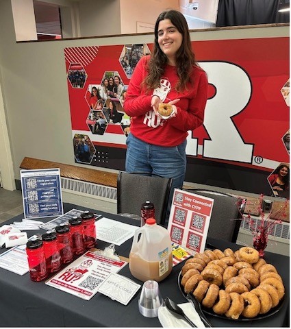 young woman with dark hair and red sweatshirt standing in front of a table set up for voter assistance and refreshments