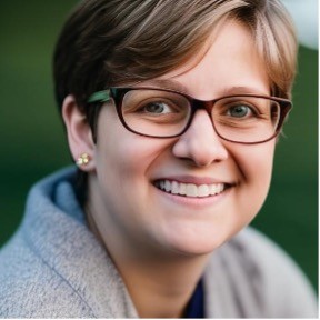 Headshot of Kristin Dulaney, a woman with pixie brown hair, brown glasses, and blue eyes smiling in front of a green backdrop.